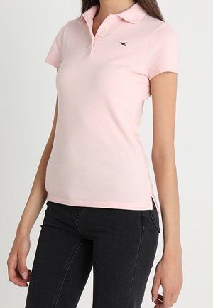 Różowa koszulka polo z krótkim rękawem, zapięta na guziki przy kołnierzyku, z wyszywanym logo na górnej lewej stronie. Noszona z czarnymi dopasowanymi dżinsami.