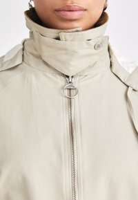 Beige jacket with high collar, zip front, and silver hardware accents. Features a textured surface and a minimalist design.