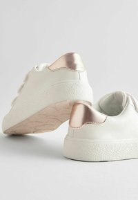Baskets blanches à la texture lisse, avec des accents métalliques roses au niveau du talon et une semelle en caoutchouc blanche à motifs. Équipées de deux bandes Velcro.