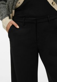 Svart skräddarsydda byxor med en slät yta, med sidofickor och bälteshällor. Kombination med en svart topp och en mönstrad cardigan.