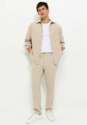 Giovane uomo che indossa una camicia e pantaloni beige con sneaker bianche, in piedi con le mani in tasca davanti a uno sfondo chiaro uniforme.