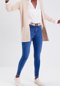 Beige long cardigan with pockets over a white blouse, blue skinny jeans, and light-colored lace-up shoes. Casual outfit with minimal details.