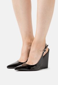 Donna con tacchi a zeppa neri in vernice, con punta affusolata e cinturini slingback, mostrata dalla parte del polpaccio in giacca bianca.