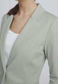 Jasnozielona marynarka z klapami z wycięciami, gładka w dotyku i o dopasowanym kroju. Noszona na białej prążkowanej bluzce, z dopasowanymi szwami na ramionach.