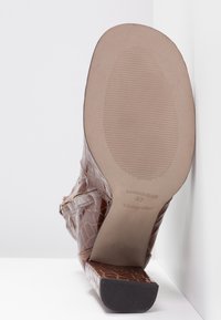 Bottine marron à talon avec motif crocodile vue de la semelle, montrant le dessous texturé et le talon bloc avec les marquages de la marque et de la taille.