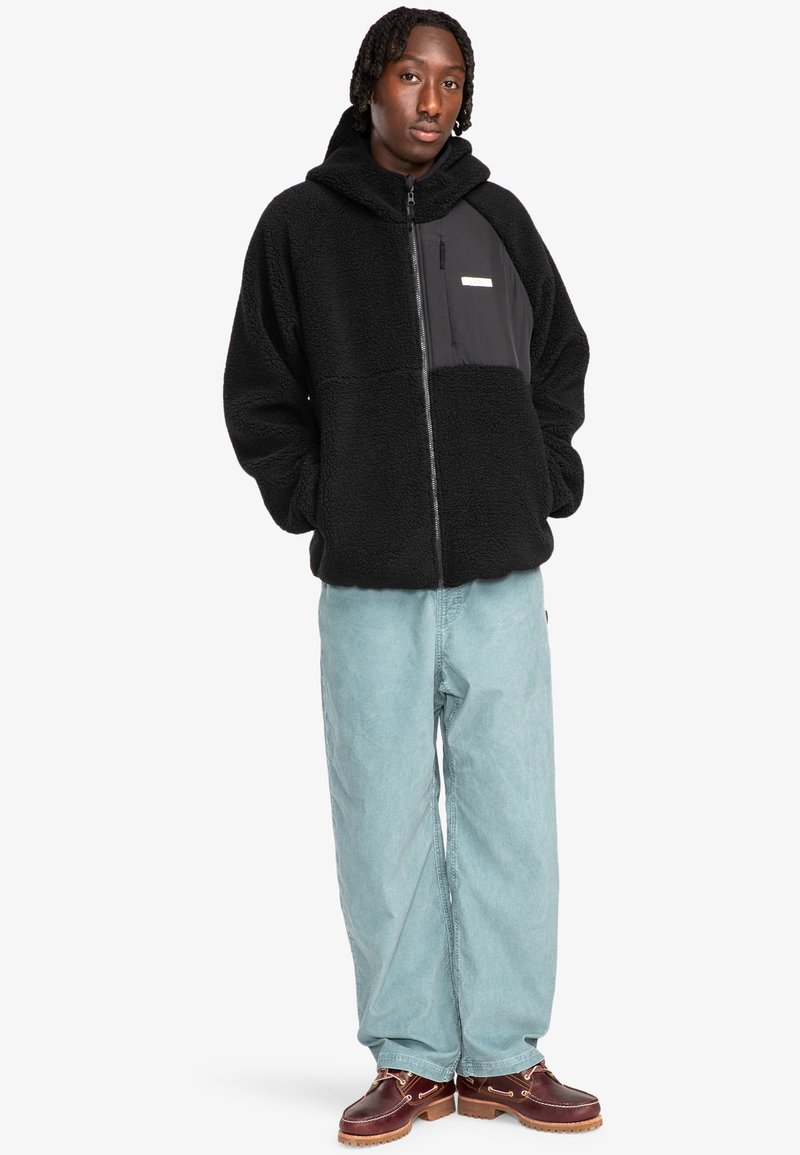 Felpa nera in pile con zip, caratterizzata da un pannello frontale liscio e nero. Abbinata a pantaloni ampi azzurri e zoccoli marroni.