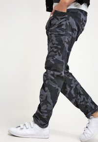 Person som bär svarta och gråa kamouflage cargo-byxor och vita velcro-sneakers som går mot en enkel bakgrund.