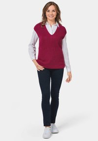 Bordeaux V-hals gebreide trui over een wit overhemd met kraag, gecombineerd met donkere jeans en witte sneakers. Gladde textuur, ontspannen pasvorm.
