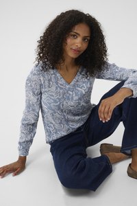 Bluse mit langen Ärmeln in Blau und V-Ausschnitt, mit einem grünen Blattmuster in Weiß. Kombiniert mit weiten, dunkelblauen Jeans und braunen Schuhen.
