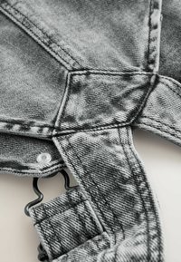 Tejido de mezclilla gris con una superficie texturizada, que presenta un botón y un detalle de clip negro para ajuste regulable. Las líneas de costura visibles realzan el diseño.