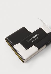 Schwarzes und weißes Lederportemonnaie mit glatter Textur, ausgestattet mit einem gelben Innenfutter und einer erhabenen Logo-Plakette, auf der "Kate Spade New York" steht.
