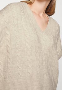 Pull en maille beige clair avec un col en V, présentant un motif en torsades, des manches courtes larges et des bords-côtes aux manches et à l'ourlet.