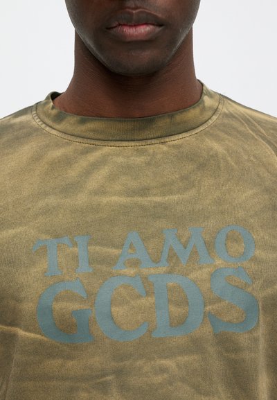 Maslinasto zelena majica s mekim tekstilom, s grafičkim tekstom svijetloplave boje "TI AMO GCDS" istaknuto prikazanim na prednjem dijelu. Okrugli dekolte.