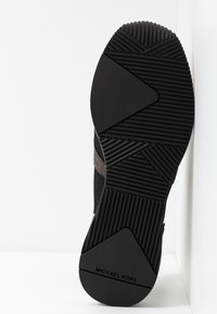 Semelle d'une sneaker noire Michael Kors avec des motifs de grip texturés et des formes géométriques, photographiée sur un fond blanc.