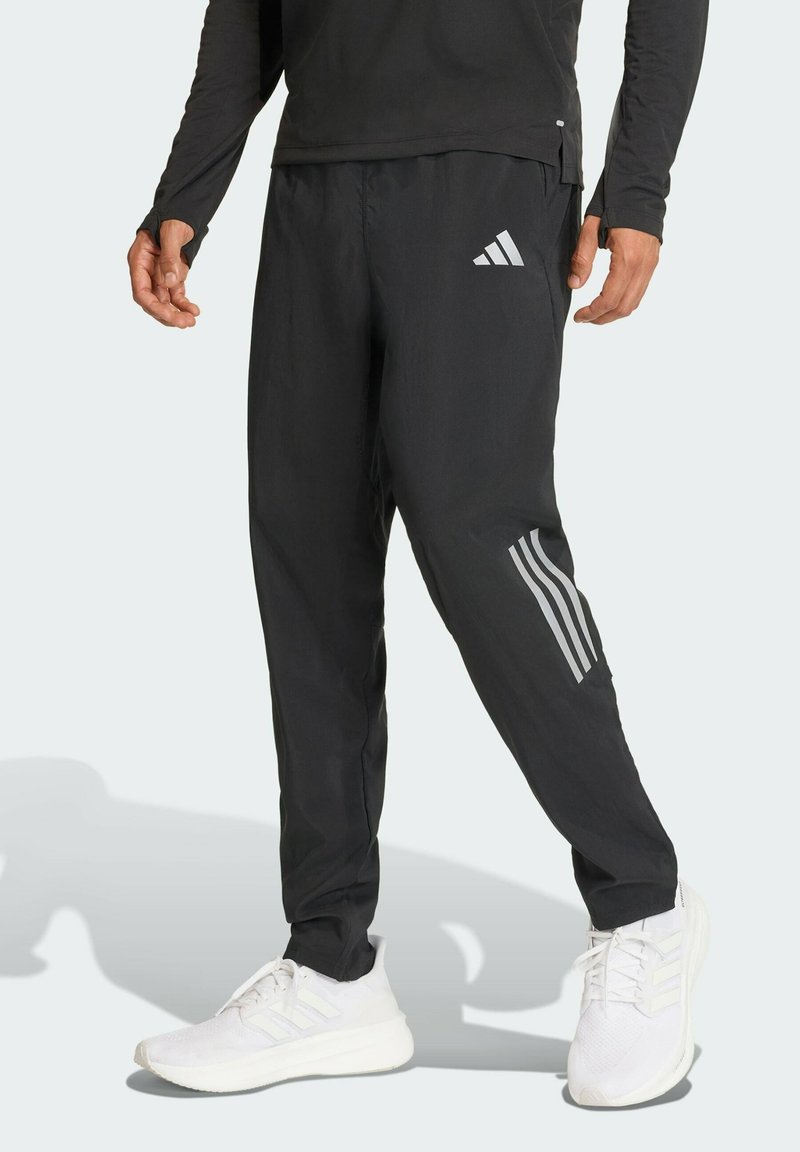 Sorte atletiske bukser med en tætsiddende pasform, adidas-logo i sølv og tre sølvstriber, der løber ned ad siden. Båret med hvide sneakers.