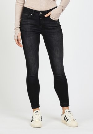 Schwarze schmale Jeans mit fünf Taschen, Knopfverschluss und leichter Ausbleichung. Kombiniert mit weißen Sportschuhen mit schwarzen Streifen.