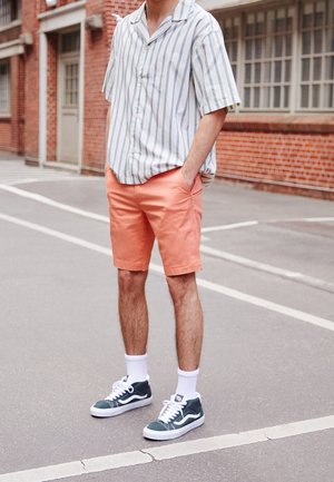 Camisa de manga corta y botones con rayas azules y blancas, combinada con pantalones cortos color coral y zapatillas verdes con suelas blancas y calcetines.
