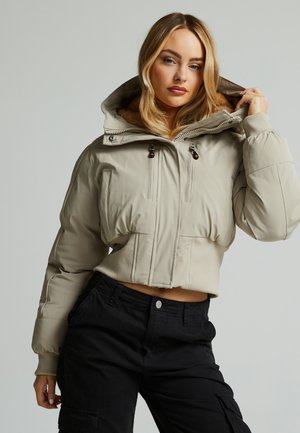 Korte beige pufferjack met capuchon, met voorzakken, ritsen en een gestructureerde stof. Gecombineerd met zwarte cargobroek.