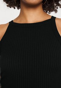 Femme portant un haut noir à côtes sans manches et des boucles d'oreilles en or, mettant en valeur les épaules et le haut de la poitrine avec des cheveux bouclés.