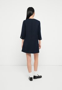 Mørkeblå kjole med trekvart ermer, teksturert stoff, glidelås bak og gullknapper på ermer.