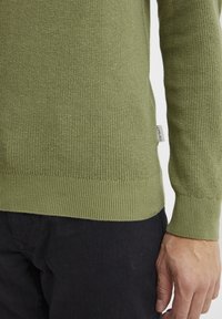 Grüner gestrickter Pullover mit gerippten Bündchen und Saum. Verfügt über ein strukturiertes Gewebe und ein kleines Markenschild an der Seite. Getragen über dunklem Stoff.