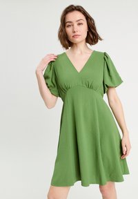 Robe verte à manches courtes avec un décolleté en V, présentant des détails froncés au niveau de la poitrine et une coupe évasée fluide, fabriquée à partir d'un tissu doux.