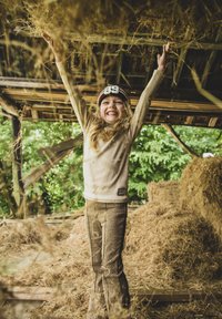 Kind met een beige lange mouw shirt en een afgedrukte bruine broek, glimlachend met opgetrokken armen in een schuur vol met hooi. Groene bladeren zichtbaar.