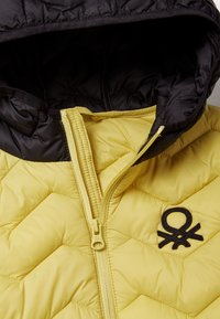 Giacca trapuntata con corpo giallo e cappuccio nero. Presenta chiusura con zip, dettaglio del logo sul lato sinistro del petto e texture liscia.