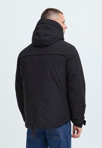 Man wearing a black hooded jacket and blue jeans, facing away against a plain light background.