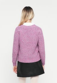 Kvinne med langt ombre-hår iført en rosa strikkegenser, hvit skjorte med krage, svart skjørt og hvite strømpebukser vendt bort mot en hvit bakgrunn.