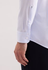 Jasnoniebieska i biała koszula w kratkę z guzikami na mankietach, gładką fakturą oraz haftem kwiatowym na mankiecie.