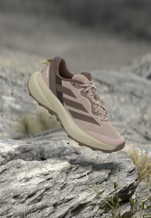 Chaussure de trail running Adidas Terrex marron clair et beige en l'air au-dessus d'un terrain rocheux avec de petites plantes, montrant une semelle robuste et un mesh respirant.