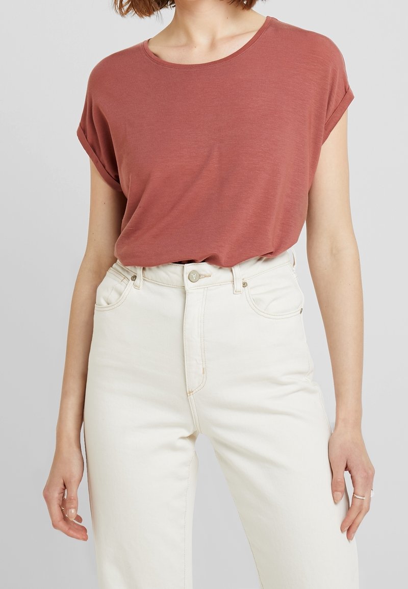 Kobieta w krótkim rękawie, w rdzawym topie, włożonym w wysokie, białe dżinsy, z rękami swobodnie opuszczonymi po bokach.