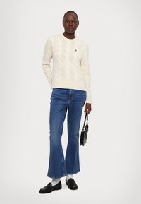 Pulover din tricot crem cu model, blugi blue flared, mocasini negri și o geantă mică neagră. Ansamblul are o croială lejeră și un design clasic.