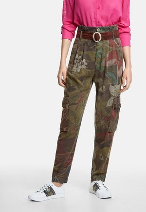 Vrouw draagt bloemmotief camouflage cargobroek met een rode riem, een roze shirt met lange mouwen en witte sneakers met patroonstrepen.