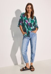 Blusa multicolore con motivi verdi, viola e neri, abbinata a jeans in denim chiaro e sandali neri con fibbie dorate.
