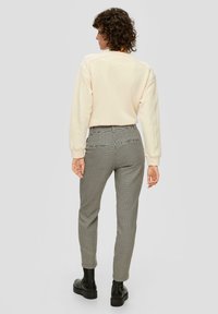 Persoon met krullend haar die met de rug naar de camera staat, draagt een crèmekleurige trui, zwarte en witte houndstooth broek en zwarte enkellaarsjes op een effen achtergrond.