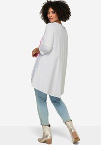 Weißes langärmliges Tunika-Oberteil mit Rüschensaum und asymmetrischem Saum, kombiniert mit hellblauen Jeans und silbernen Ankle Boots. Einfaches Design ohne Muster.