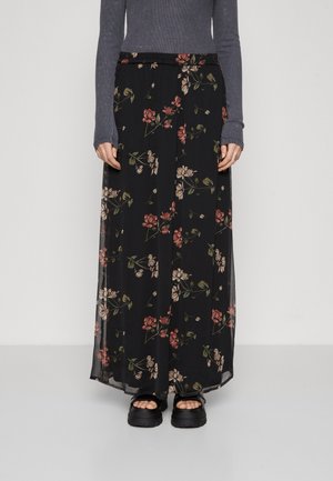 Femme portant une jupe longue noire transparente à fleurs associée à des sandales noires à plateforme et un haut gris ajusté à manches longues.