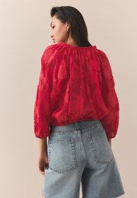 Červená priesvitná blúzka s textúrovaným kvetinovým vzorom, zošitý výstrih a voľné rukávy, doplnená modrými denimovými šortkami s vysokým pásom.