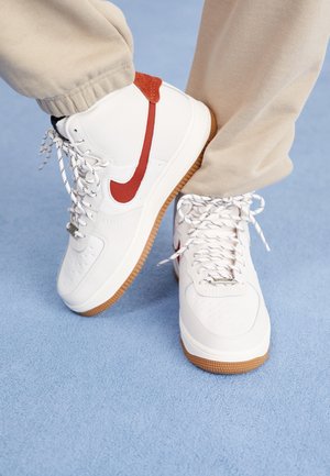 Baskets montantes blanches avec le logo Nike rouge et semelles gomme, portées avec un pantalon beige sur un sol texturé bleu clair.