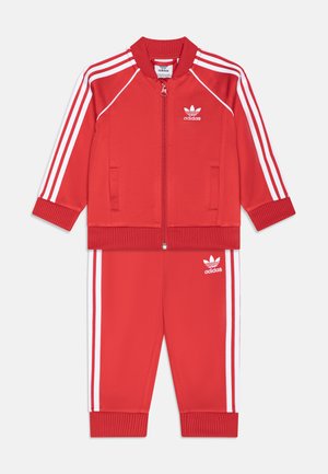 Tuta Adidas rossa caratterizzato da una giacca con zip con strisce bianche e pantaloni abbinati. Realizzato in un tessuto liscio e strutturato. Accenti del logo bianco.