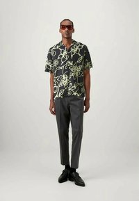 Svart kortärmad skjorta med grönt blommönster, kombinerad med grå skräddarsydda byxor och svarta skor. Modellen har också på sig mörka solglasögon.