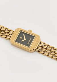 Zlaté kovové náramkové hodinky so štvorcovým, vzorovaným puzdrom, čiernym ciferníkom a zlatým náramkom. Obsahujú malé okno na zobrazenie dátumu a dve ručičky.