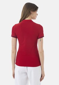 Rød polo-shirt med korte ærmer, der har blå detaljer på ærmerne. Stoffet ser glat og strækbart ud, med et slim fit-design.