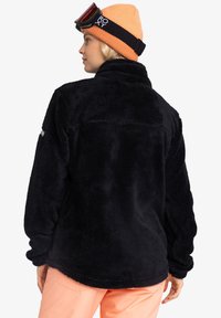 Černý fleece svetr s vysokým límcem, texturovaným povrchem a dlouhými rukávy. Obsahuje zadní kapsu a je doplněn jasně oranžovou čepicí.