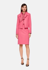 Rosa taillierte Jacke und Bleistiftrock-Set, mit einer Bluse mit Schleife darunter. Das Material scheint strukturiert zu sein, hat eine körperbetonte Silhouette und eine lebendige Farbe.