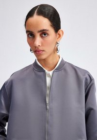 Grå satin bomberjacka med dragkedja fram, ribbad krage och sidofickor, bärs över en vit skjorta med krage. Silverfärgade droppformade örhängen.