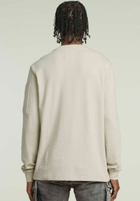 Kremowy długi sweter z teksturowanej tkaniny. Posiada okrągły dekolt i dopasowane mankiety. Gładki tył z kieszonką na lewym rękawie.