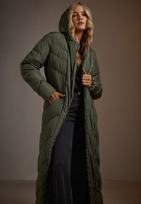 Długi, ocieplany płaszcz w ciemnozielonym kolorze z kapturem, o pikowanej strukturze i bocznymi kieszeniami, rozpięty, odsłaniający czarny top pod spodem.
